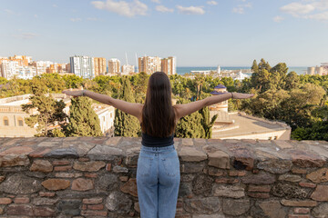 Fototapeta premium Woman with long hair stands on stone wall, arms outstretched, overlooking cityscape and ocean, embracing freedom and nature's beauty