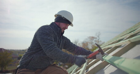 Construction worker hammering wooden beams on roof, wearing helmet and gloves, focus on labor and craftsmanship in rooftop framing, residential building project