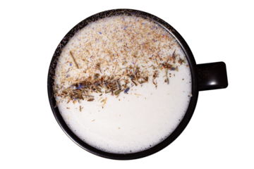 A black ceramic cup filled with thick milk foam, topped with a horizontal stripe of dried herbs and flower petals including lavender, cornflower, and meadow plants.