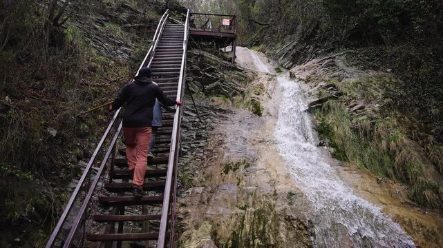 A lone adventurer ascends a rustic metal stairway next to a flowing waterfall, surrounded by forest.