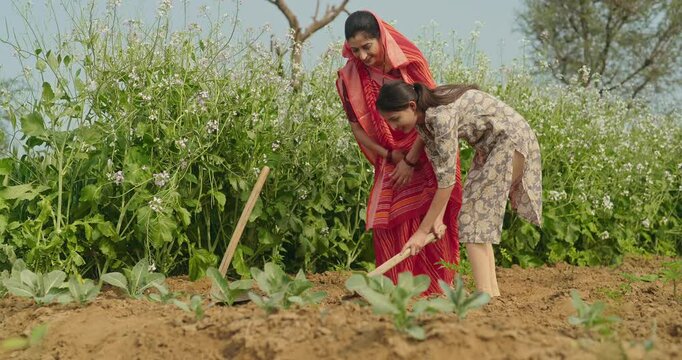 Indian happy young woman wear red saare with teen age girl talking working together hold hoe tool prepare planting at farmland asian smiling mother teach dig soil outdoor desi life