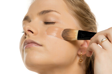 Close-up of young woman applying liquid foundation with makeup brush on cheek, showing smooth...