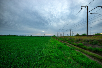 Dirt Path Running Alongside a Green Field and Railway Tracks Under a Cloudy Dawn
