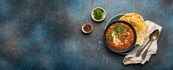 Traditional Indian Punjabi dish Dal makhani with lentils and beans in black bowl served with naan flat bread, fresh cilantro and two spoons on blue concrete rustic table top view. Space for text.