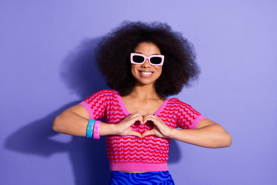 Stylish woman with striped top and retro hairstyle making a heart gesture against a vibrant purple background