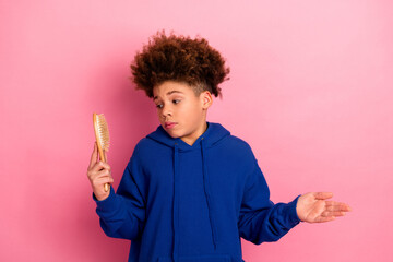 Young boy in blue hoodie holding a hairbrush, expressing playful uncertainty against a pink...
