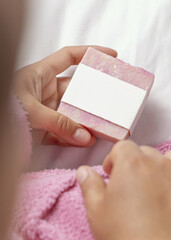 Closeup of woman in pink bathrobe holding soap bar with blank label over white bedsheet, mockup