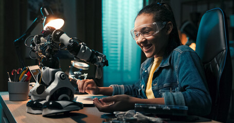 Portrait of a smiling girl checking toy repair instructions on her laptop. She scrolls through web pages and pulls electronic cables from a robot, focused on fixing it herself.