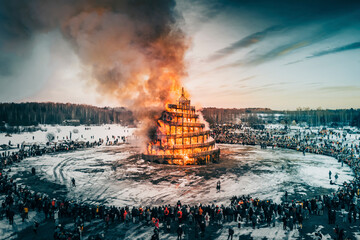 Aerial view of a festive winter celebration with a large bonfire and crowd amidst snow-covered forest, Ugorskoe, Kaluga, Russia.