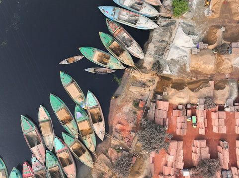 Aerial view of colorful traditional boats in a serene river with a rustic village and urban architecture, Dhaka, Dhaka, Bangladesh.