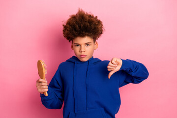 Young boy in blue hoodie expressing disapproval with thumbs down gesture holding hairbrush on pink...