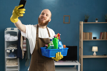 Young bearded man wearing gloves and apron holding basket with cleaning supplies taking self...