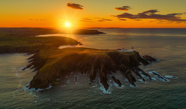 Aerial view of dramatic sunset over rugged cliffs and serene ocean with a picturesque lighthouse, Rathbarry, County Cork, Ireland.