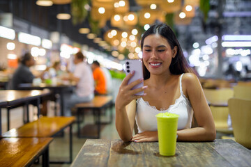 Young woman using phone and texting while drinking healthy avocado smoothie drink in cafe at night