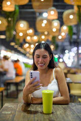 Young woman using phone and texting while drinking healthy avocado smoothie drink in cafe at night