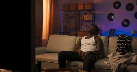 Obraz premium A sporty male with a multi-racial background sits on a couch in a white strapped T-shirt, looking away in silence, expressing deep thought and subtle disappointment.