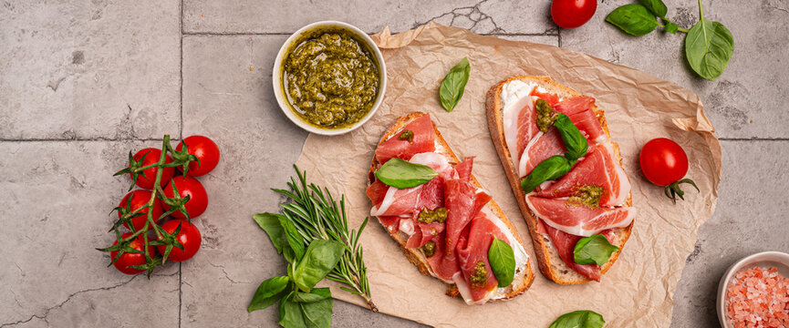 Grilled toast with jamon or prosciutto, cream cheese, pesto and basil on a paper on gray background for breakfast top view. Healthy homemade breakfast or lunch idea.