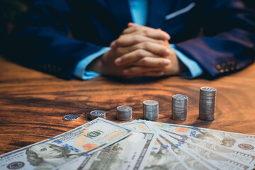 Businessman in Suit with Hands Folded over Table Surrounded by Coins and Dollar Bills Currency...