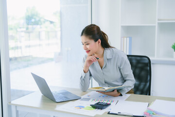 Asian businesswoman using laptop while analyzing report in bright modern office.