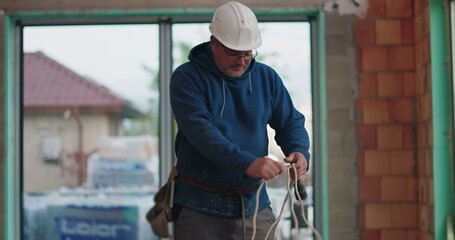 Construction worker in blue hoodie organizing ropes, hard hat, on-site, indoor construction setup, preparing tools, focus on task in building renovation