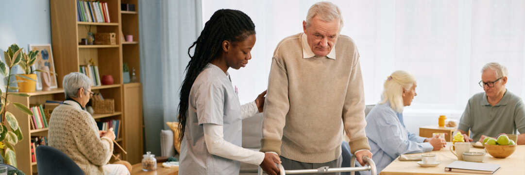 Caregiver helping elderly man using walker in cozy care home with other residents engaged in activities and mealtime in the background