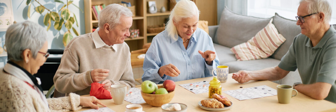 Group of elderly friends sitting around a table, engaged in playing bingo and sharing snacks. Senior individuals smiling and interacting, seen in bright and homely environment - Powered by Adobe
