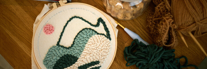 Detailed view of an ongoing embroidery project, showcasing a landscape pattern and yarn organized around Embroidery hoop placed on wooden table, highlighting hand craftsmanship