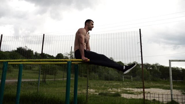 Strong man trains abdominal muscles raising legs on parallel bars at sports ground. Young shirtless sportsman exercising at nature. Sporty athlete working out at nature. Concept of sportive lifestyle