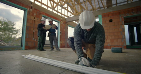 Construction worker kneeling and measuring PVC pipes in unfinished building interior, with...
