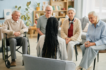 Caregiver with long braids addressing group of seniors seated in cozy living room setting, engagement apparent as all seniors focus attentively on caregiver speaking