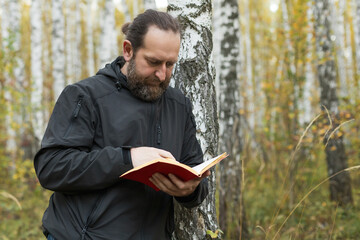A man with a beard is reading a book in a forest