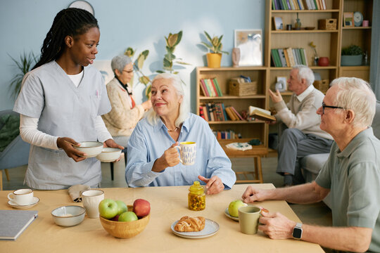 Caregiver serving tea and snacks to seniors in cozy community room filled with books and plants. Seniors engaged in various activities meeting physical and social needs