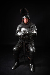 Portrait of a knight in full plate armor holding a sword posing sitting on chair isolated on a dark...