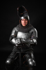 Portrait of a knight in full plate armor holding a sword posing sitting on chair isolated on a dark...