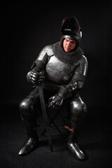 Portrait of a knight in full plate armor holding a sword posing sitting on chair isolated on a dark...