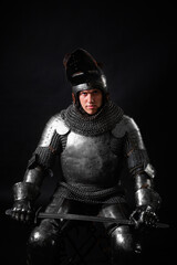Portrait of a knight in full plate armor holding a sword posing sitting on chair isolated on a dark...