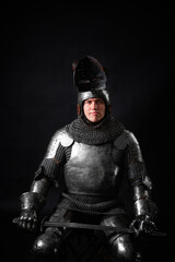 Portrait of a knight in full plate armor holding a sword posing sitting on chair isolated on a dark...