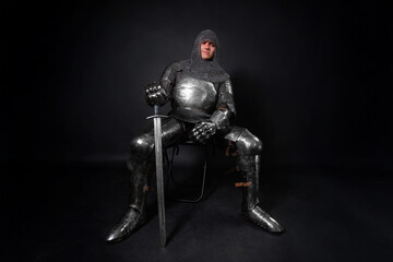 Naklejka premium Portrait of a knight in full plate armor holding a sword posing sitting on chair isolated on a dark background.