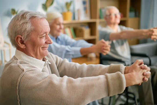 Elderly people in retirement home performing stretching exercises while seated, focusing on health and well-being amid light-filled indoor setting with plants and bookshelves - Powered by Adobe