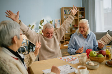 Group of elderly friends enjoying each other's company while playing a board game around dining table, with cheerful expressions and raised hands in celebration