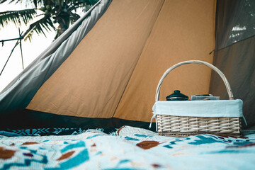 A cozy camping scene features a woven basket filled with cookware, draped with a white cloth inside...