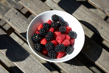 A bowl filled with red raspberries and blackberries is placed on a wooden table. The vibrant colors of the berries contrast beautifully with the natural surroundings and sunlight