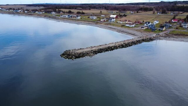 Aerial view of an old wharf protected by a wave-break rock wall that juts out into a calm saltwater bay surrounding a pretty coastal village. Baie-des-Sables, Quebec, Canada, 2025.