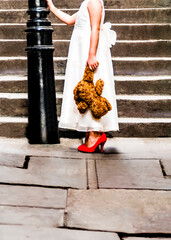 A young girl in a white dress holding a red shoes and a teddy bear 