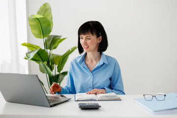 Businesswoman works on laptop at desk, with calculator, notebook, glasses nearby. Modern home or office workspace with big indoor plant. Concept of remote work, freelance, online education, accounting