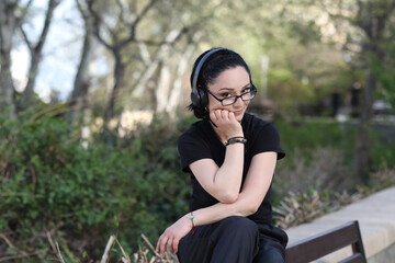 Confident young woman with glasses and headphones sitting on a bench in a park, resting her chin on her hand and smiling slightly, enjoying a peaceful moment