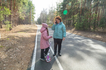 Grandmother talking on the phone while walking with her granddaughter who is riding a scooter in...
