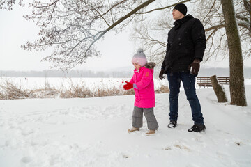 Father with daughter dressed warmly and in warm gloves builds a snowman in a snowy near the lake.