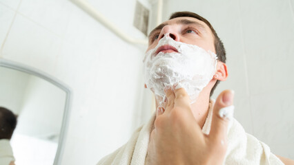 Young man applying shaving foam near mirror in bathroom