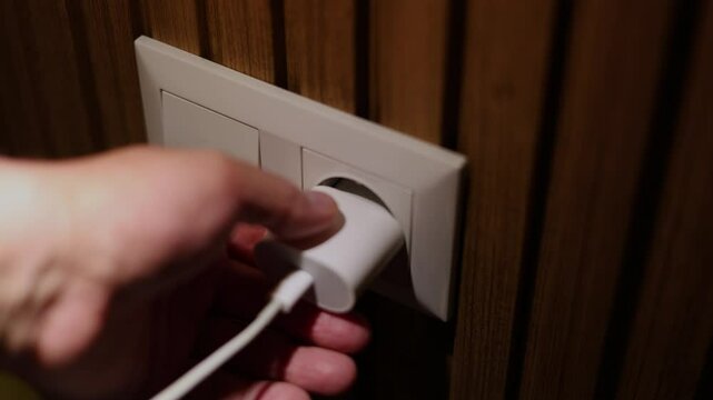 Horizontal defocused 4K video. Male hand inserts mobile phone charger plug, gadget, laptop, extension cord, computer, TV, kettle in white wall socket on wood wall in dark hotel room. Selective focus